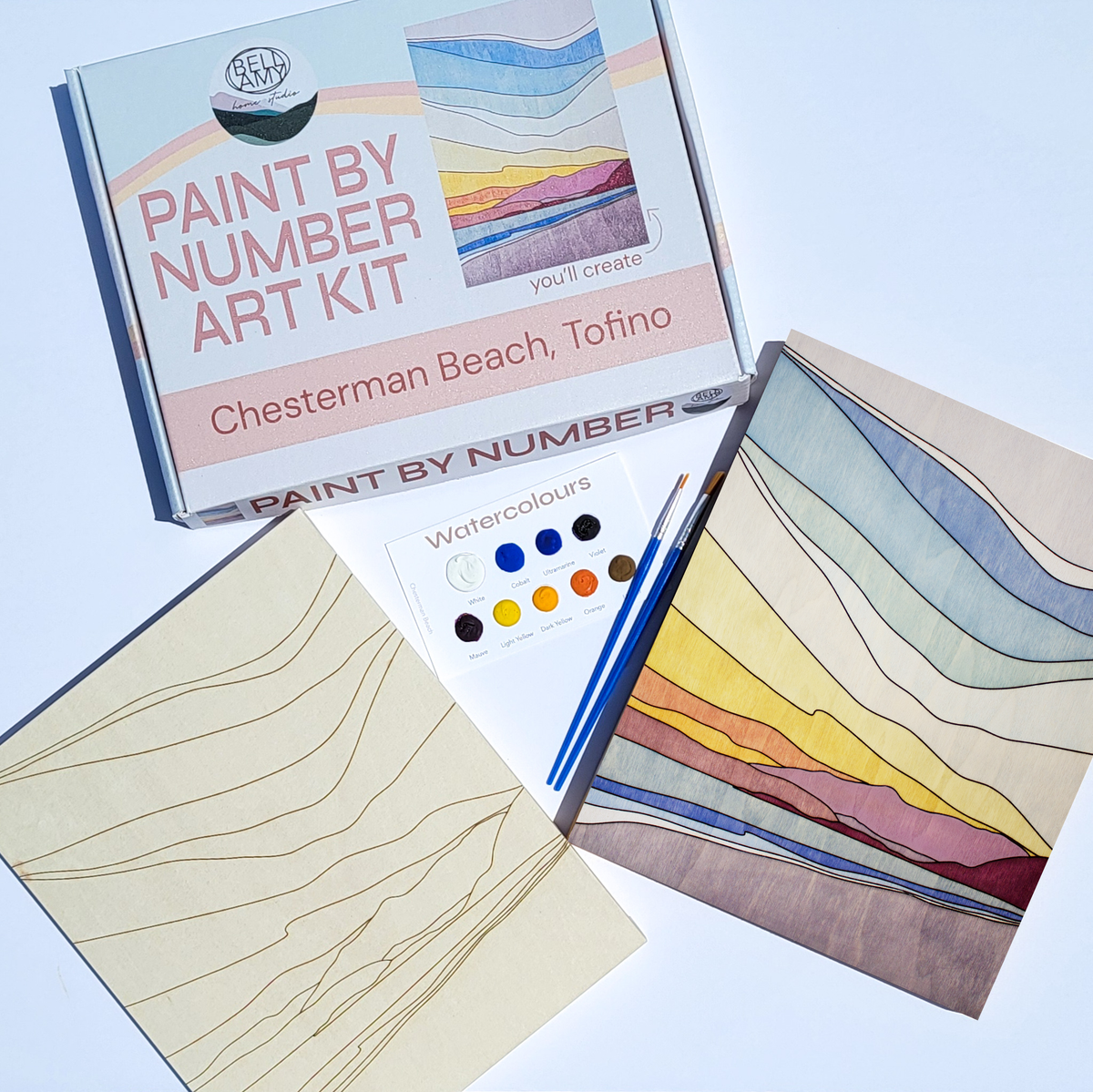 Bellamy Home Studio Paint By Number Art Kit for Chesterman Beach, Tofino — kit contents flat-laid on a light surface showing the branded box, engraved wood panel, watercolour palette in warm sunset tones, paint brushes, and a partially completed coastal landscape. Handmade in Canada.
