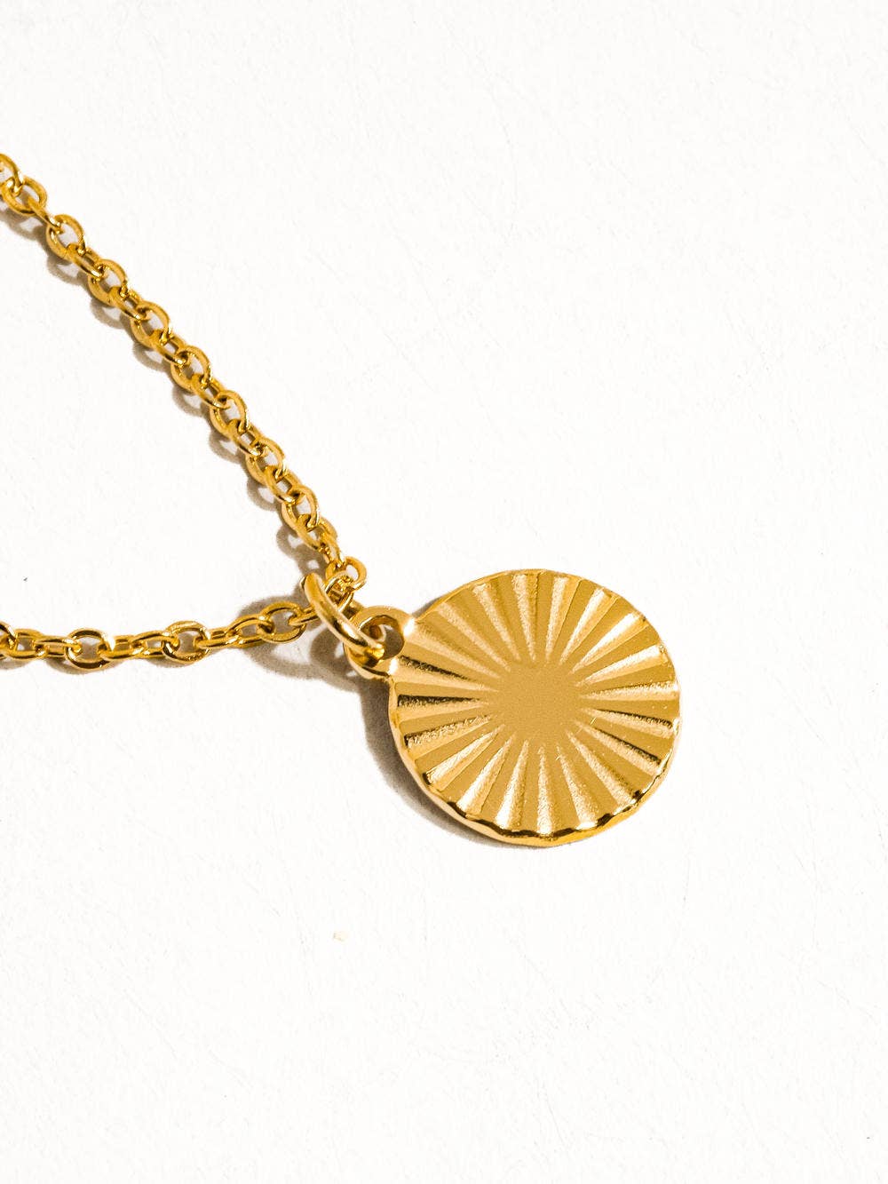 Miraz Sun Gaze Necklace by Flânerie by MAIQ with a round gold sunburst pendant and gold link chain, resting on a white background.