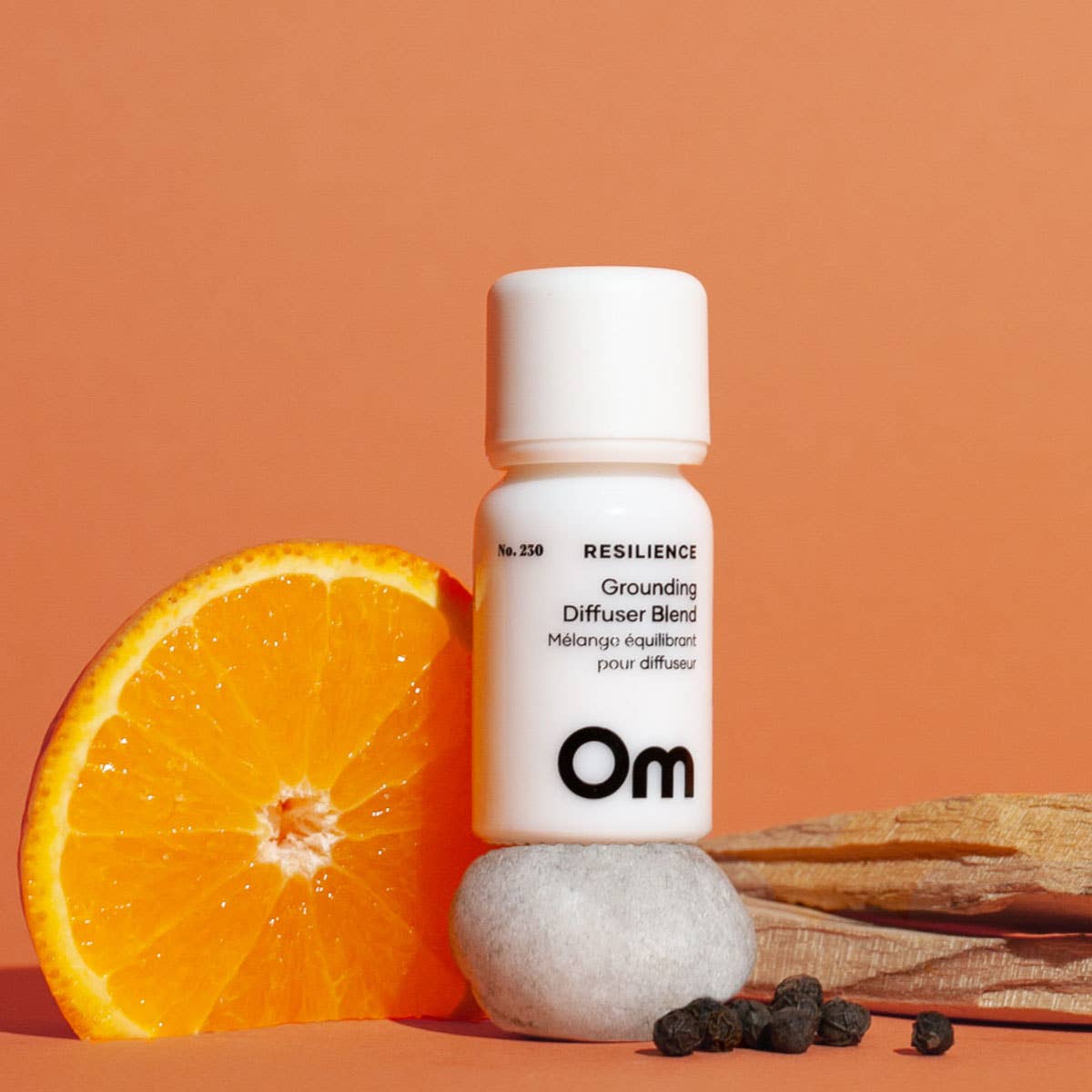 Om Resilience Grounding Diffuser Blend No. 230 in a white glass bottle, styled with fresh orange, palo santo, and black pepper on a terracotta backdrop for an earthy, calming aromatherapy moment.