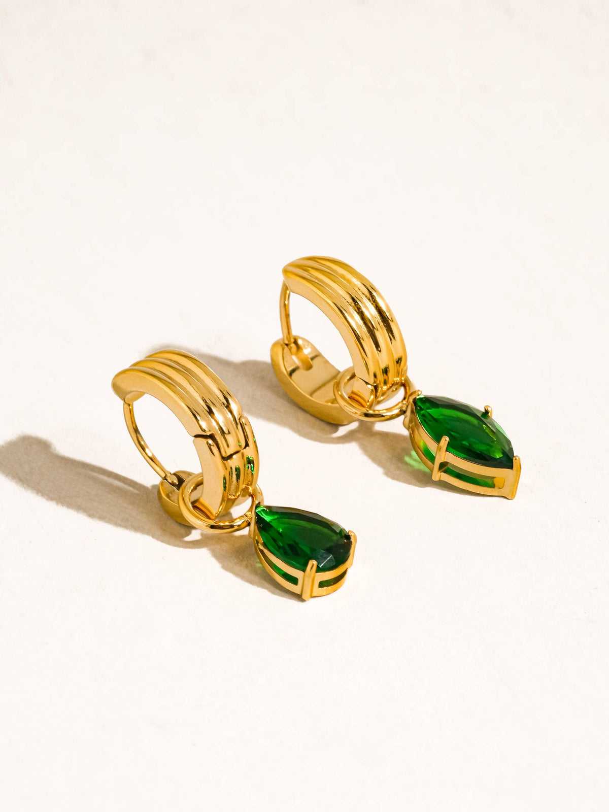 Saffron Teardrop Earrings by Flânerie by MAIQ with ridged gold hoops and deep green teardrop stones in gold settings, resting on a cream background.