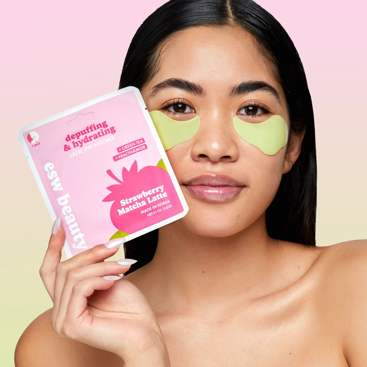 A woman with green under-eye patches holds a pink package of esw beauty Strawberry Matcha Under Eye Patches against a soft pink and yellow gradient background. K-beauty hydrating and de-puffing undereye patches.