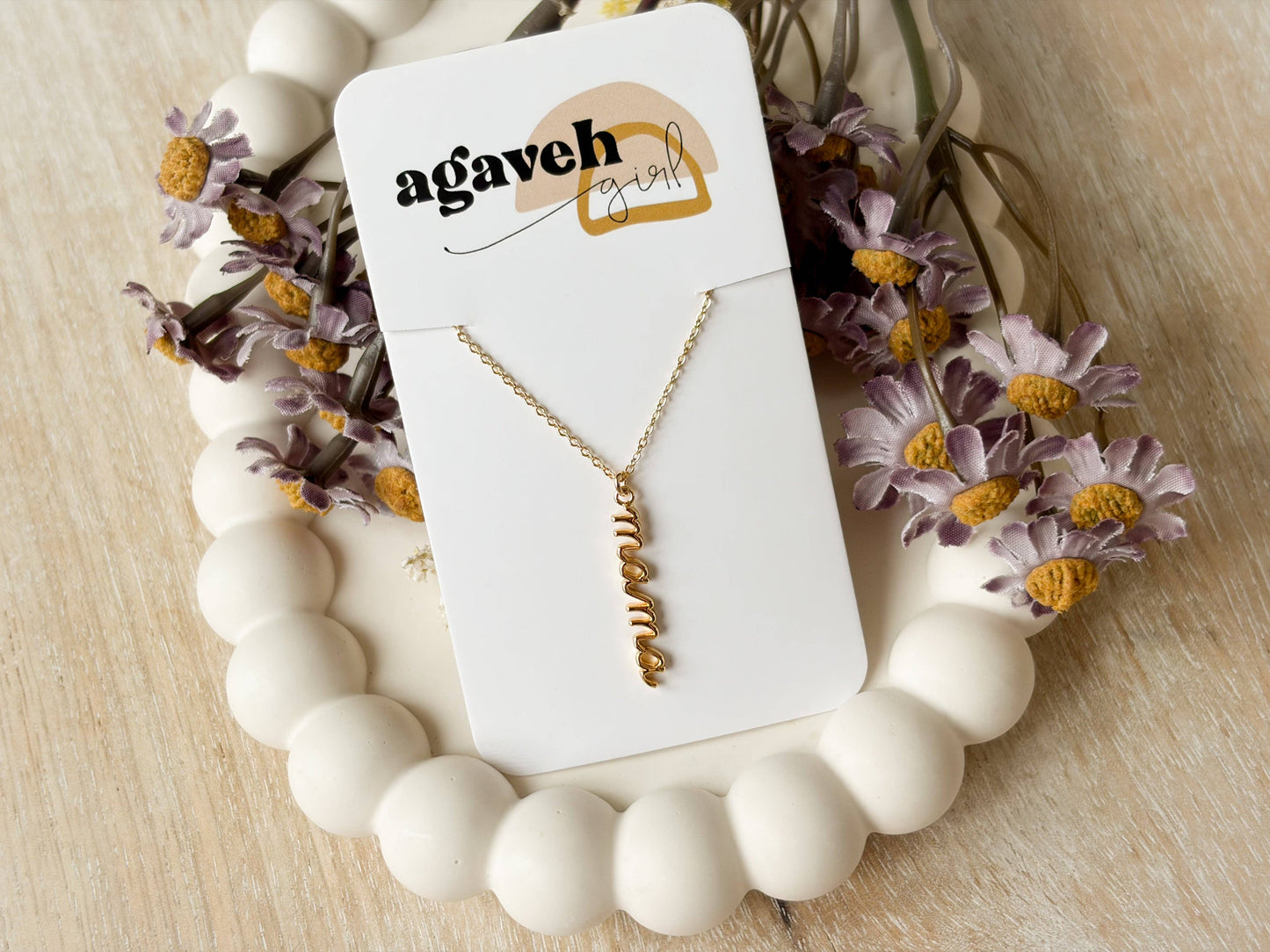 Agaveh Girl Mama Nature Necklace in gold-plated handwritten script, displayed on a branded card nestled in a white bead garland with dried purple daisy florals. Made in Canada.
