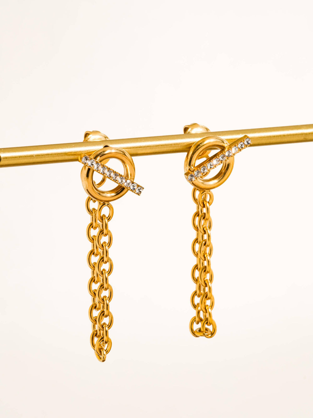 Lisle Chain Drop Earrings by Flânerie by MAIQ with gold circular studs crossed by clear stone-set bars and dangling link chains, displayed on a gold rod against a cream background.