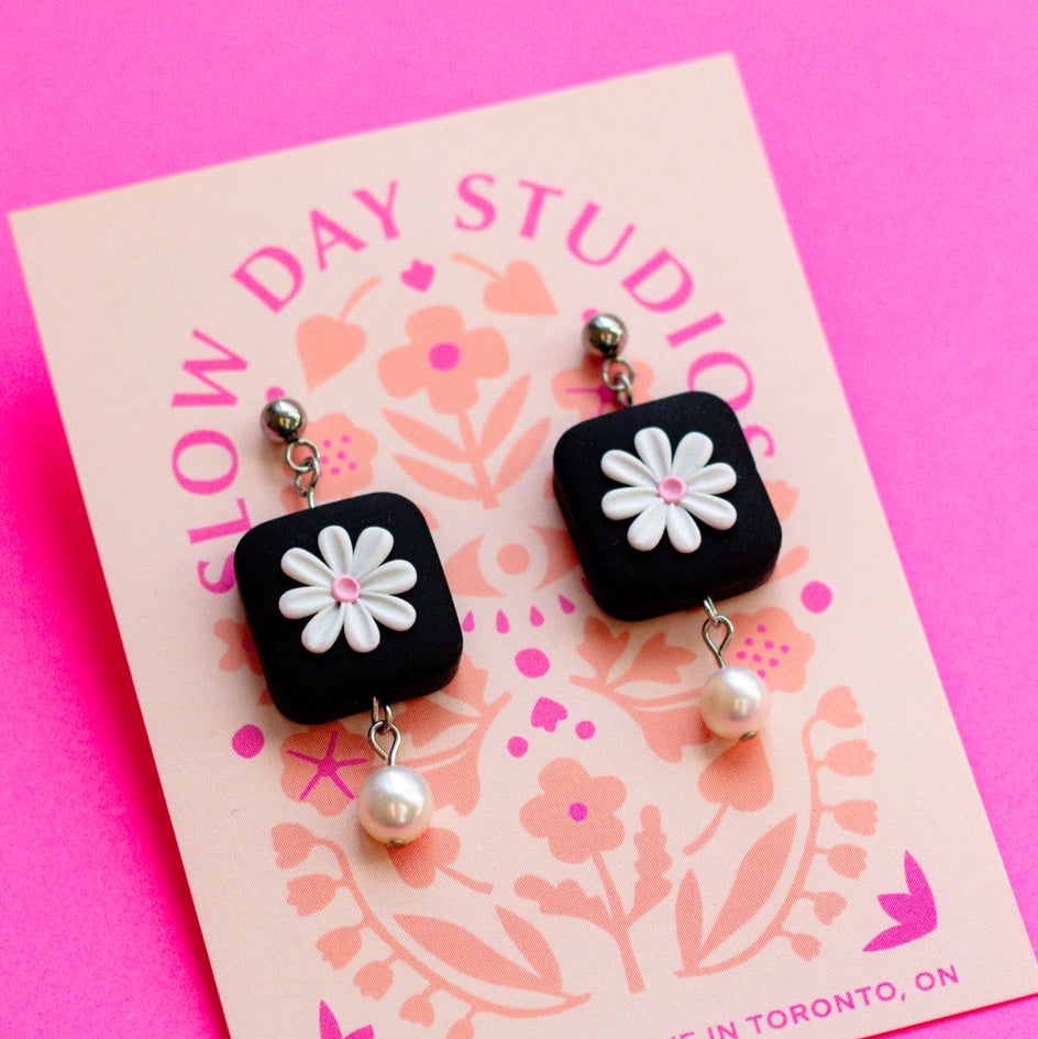 Black square earrings with white flowers and pearls on a pink card with 'Slow Day Studios' branding.