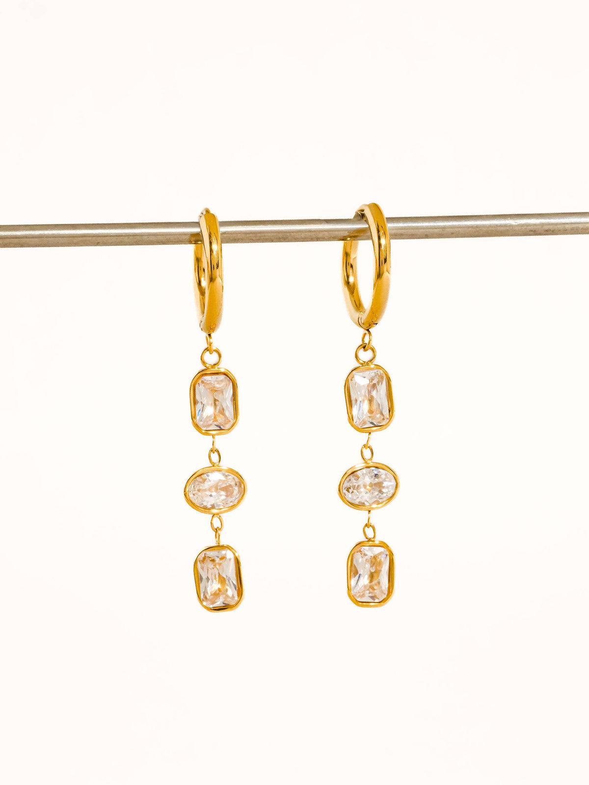 Sunniva Triple Drop Earrings by Flânerie by MAIQ with gold hoops and three clear rectangular and oval cubic zirconia stones, hanging from a metal rod against a cream background.