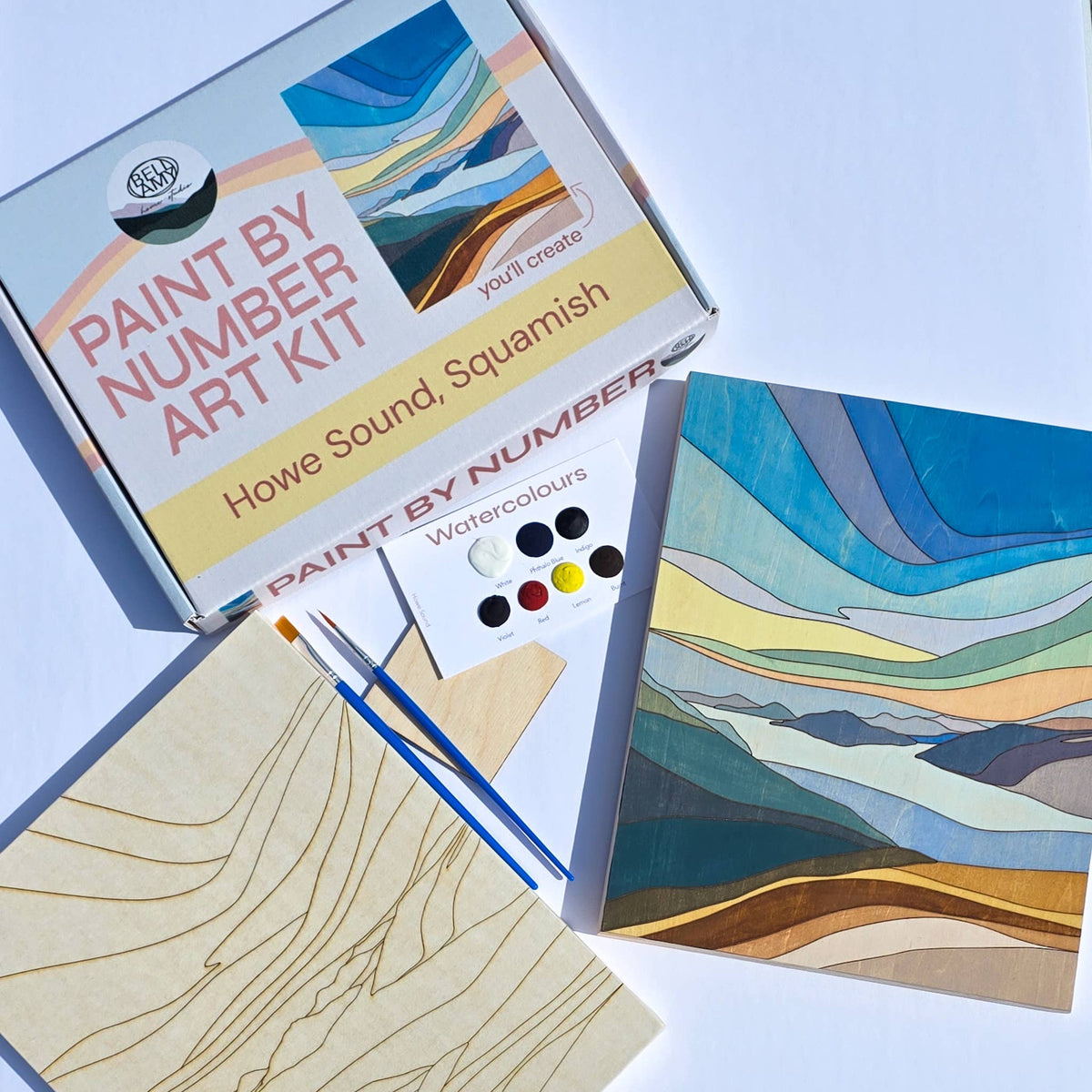 Bellamy Home Studio Paint By Number Art Kit for Howe Sound, Squamish — kit contents laid flat showing an engraved wood panel, paint brushes, watercolour palette, and a completed example of the layered mountain and water landscape scene. Handmade in Canada.