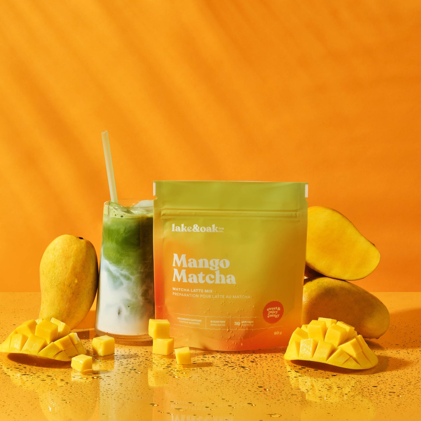 Lake & Oak Mango Matcha Latte Mix pouch surrounded by fresh mangoes and an iced matcha drink against a vibrant orange background. 
