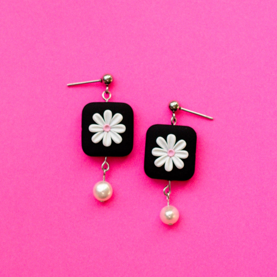 Black square earrings with white flowers and pearl drops on a pink background