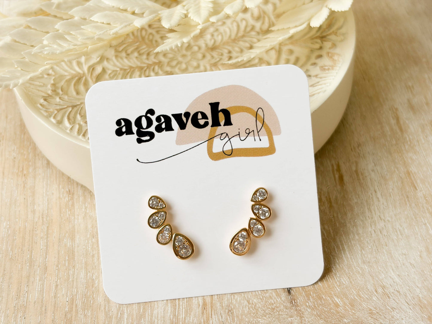 Agaveh Girl Angel Wings ear climber studs in 14K gold-plated brass with pear-shaped cubic zirconia stones, displayed on a branded card against a white decorative dish. Made in Canada