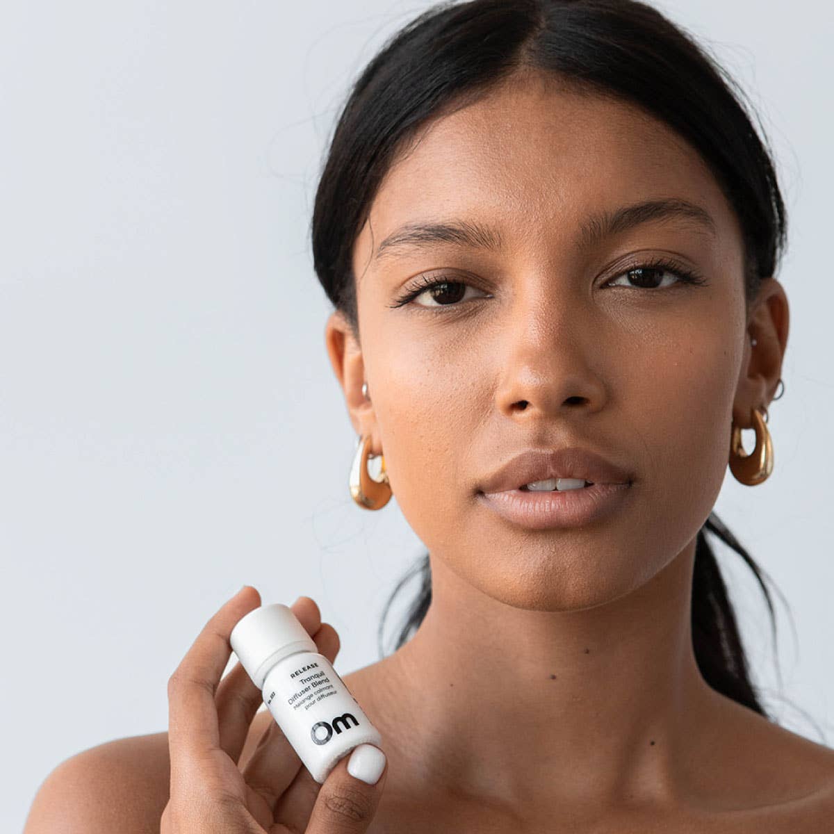 A woman holding a white cosmetic rollerball bottle with the brand 'om' visible, set against a clean light gray background.