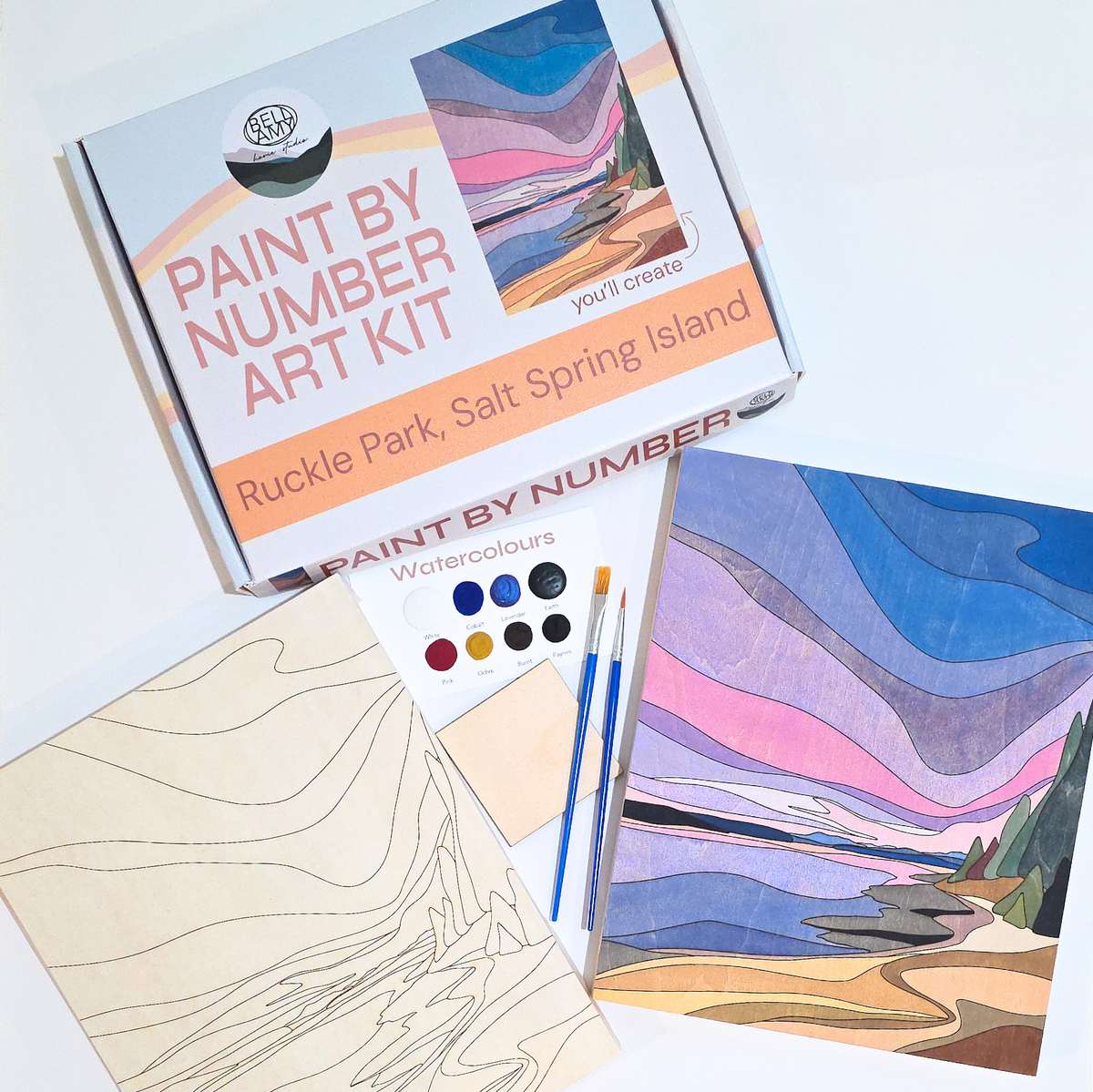 Bellamy Home Studio Paint By Number Art Kit for Ruckle Park, Salt Spring Island — kit contents spread on a white surface showing the branded box, engraved wood panel, watercolour palette, paint brushes, and a completed example of the pink and purple coastal landscape. Handmade in Canada.