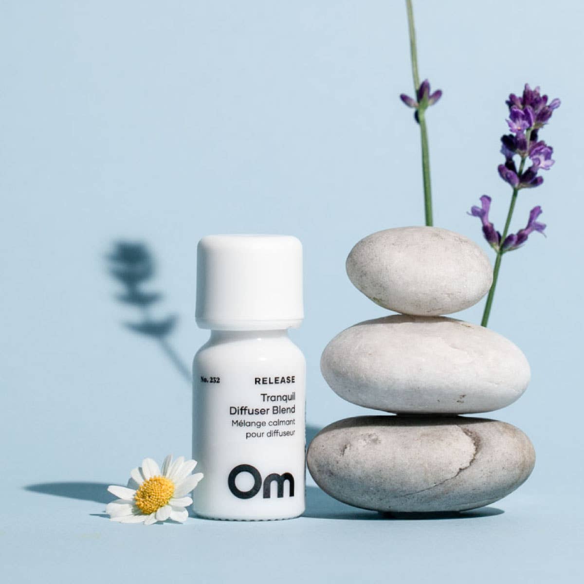 A small white bottle labeled 'Om Release Tranquill Diffuser Blend' stands beside stacked smooth stones and lavender sprigs on a pale blue background.