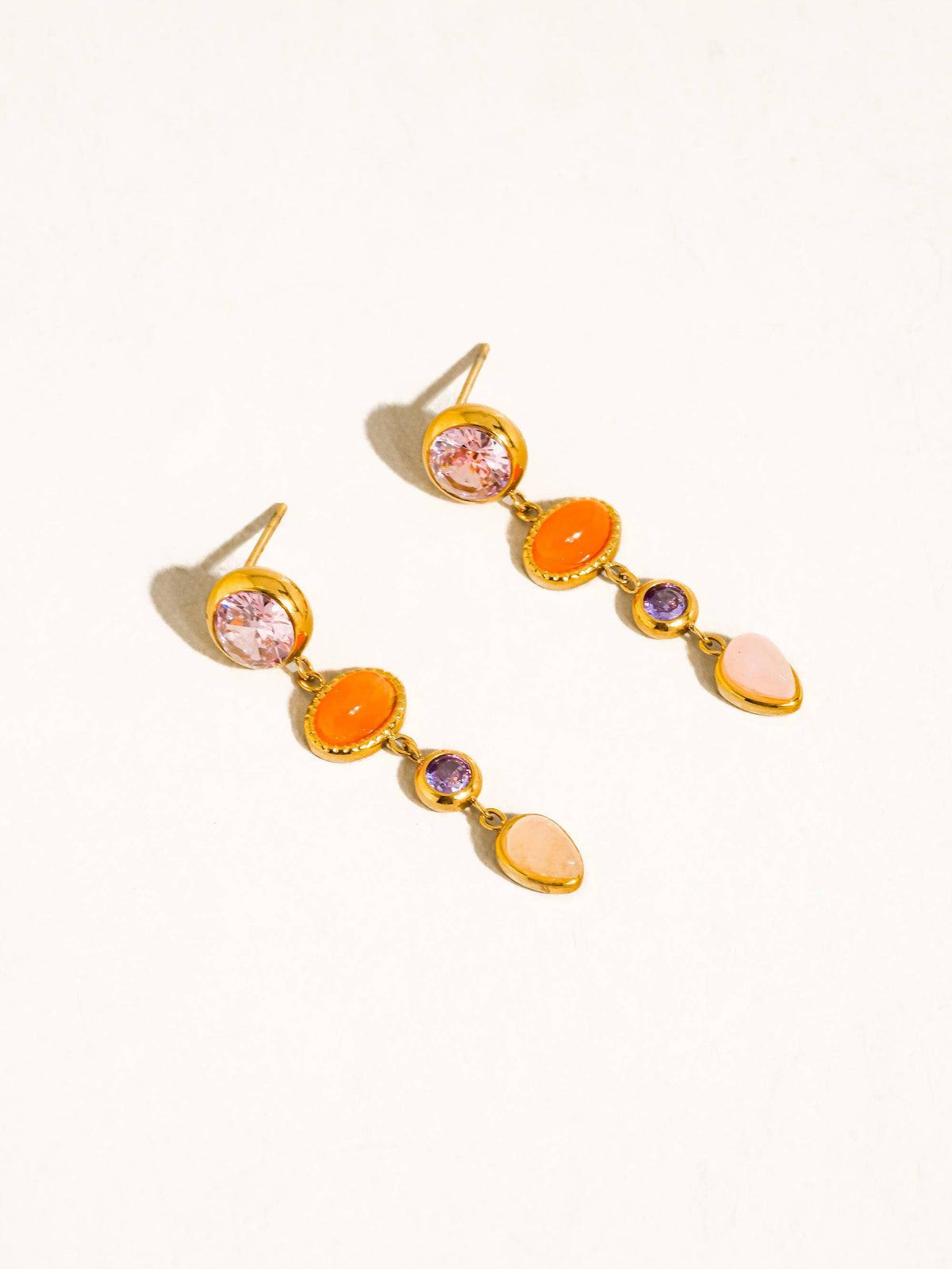 Évisa Gemstone Drop Earrings by Flânerie by MAIQ with pink, orange, purple and pale pink stones linked in gold settings, arranged on a cream background.
