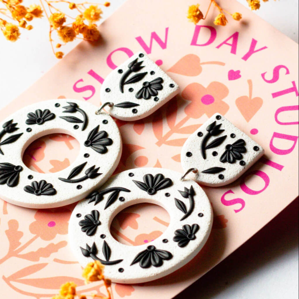Decorative earrings with black floral patterns on a pink surface with 'Slow Day Studios' branding.