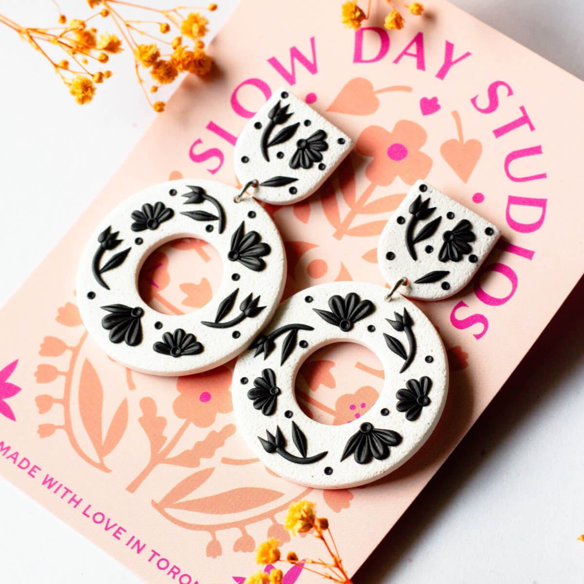 White earrings with black floral patterns on a pink 'Slow Day Studios' card.
