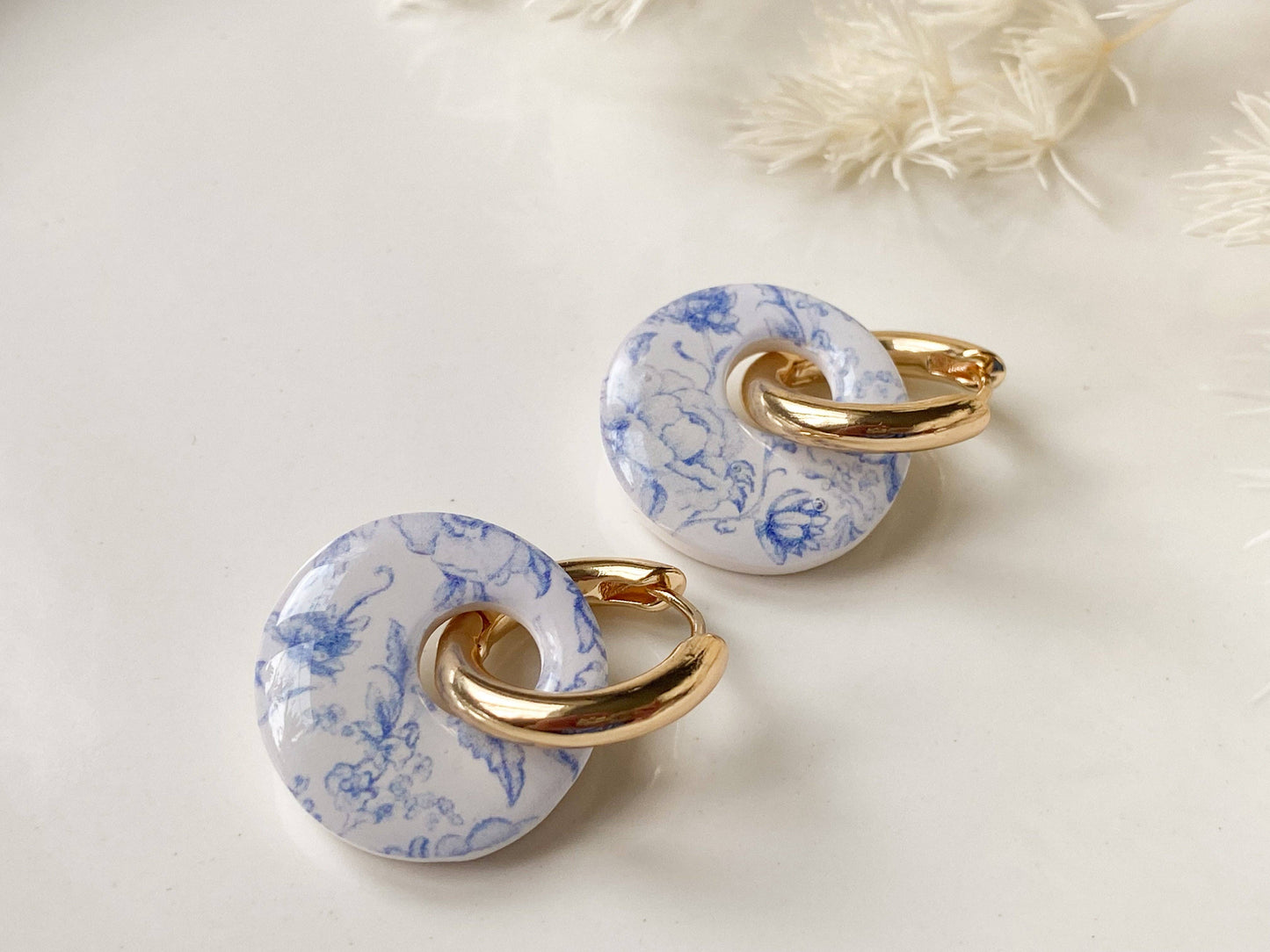 Agaveh Girl Madrid Hoops in Blue Floral, showing the gold hoop threaded through a round blue and white floral polymer clay disc, styled on a clean white surface with soft feathery texture in the background. Made in Canada.