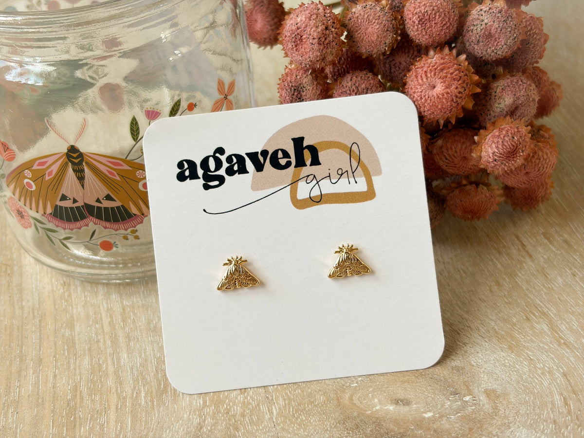 Agaveh Girl Moth Studs, made in Alberta, Canada, with detailed gold wings on a white branded card beside a moth-decorated glass jar and dried pink flowers. 