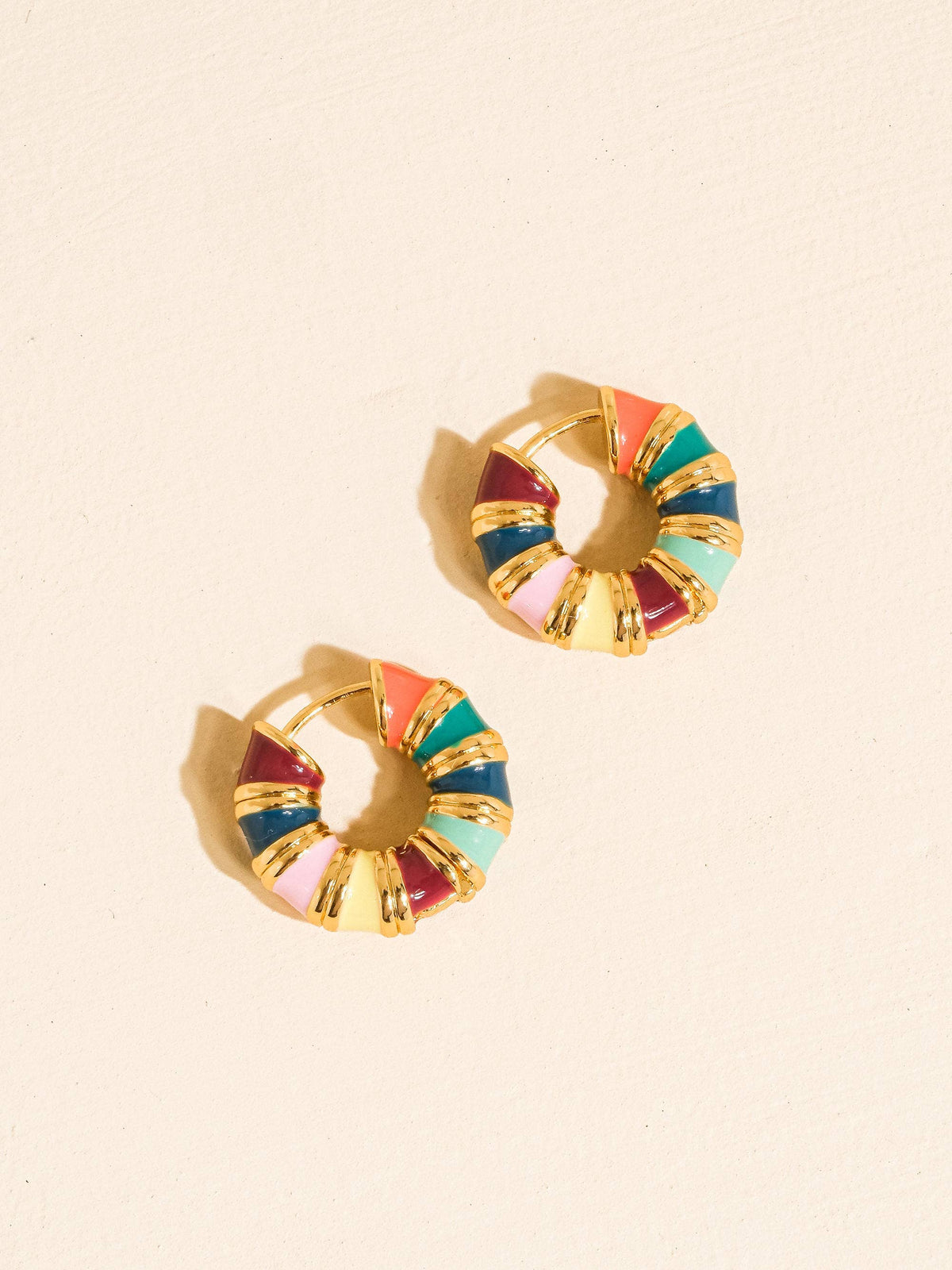 Ágata hoop earrings by Flânerie by MAIQ with multicoloured enamel segments and textured gold dividers, arranged on a peach background with soft shadows.