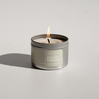 A Bonfire + Balsam scented beeswax candle tin made in Edmonton, AB. The candle has scent notes of woodsmoke, red cedar and frankincense. The candle tin is silver with a sage green label and is shown burning with a white backdrop.