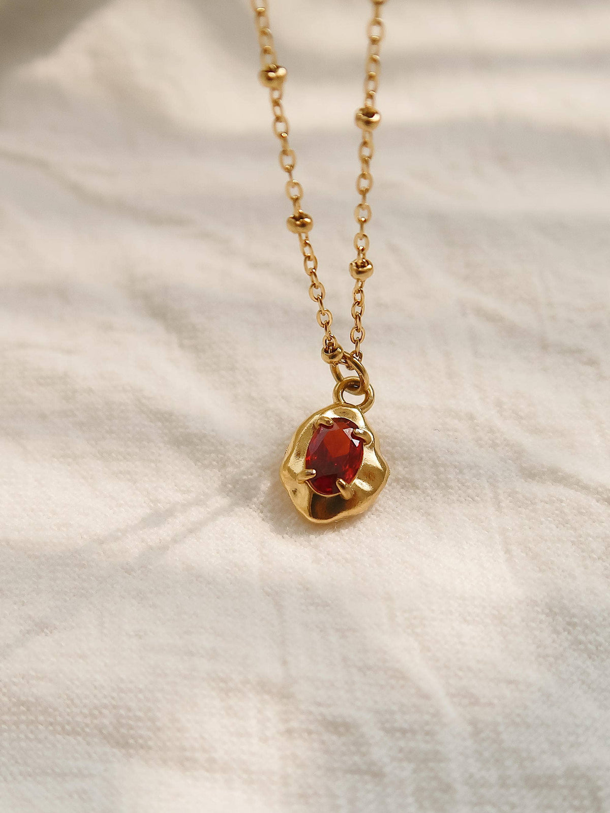 Ryleigh Petite Necklace by Flânerie by MAIQ with a red stone in an irregular gold pendant on a delicate beaded gold chain, resting on cream fabric.