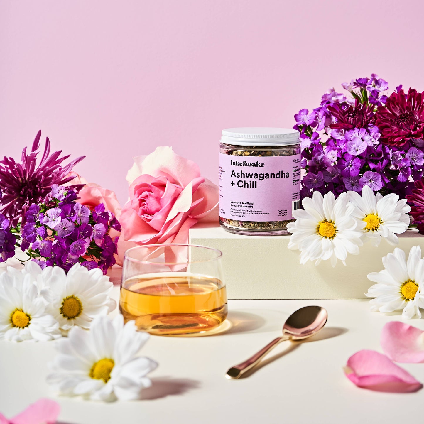 Jar of 'Ashwagandha + Chill' supplement with a glass of tea and flowers on a pink background