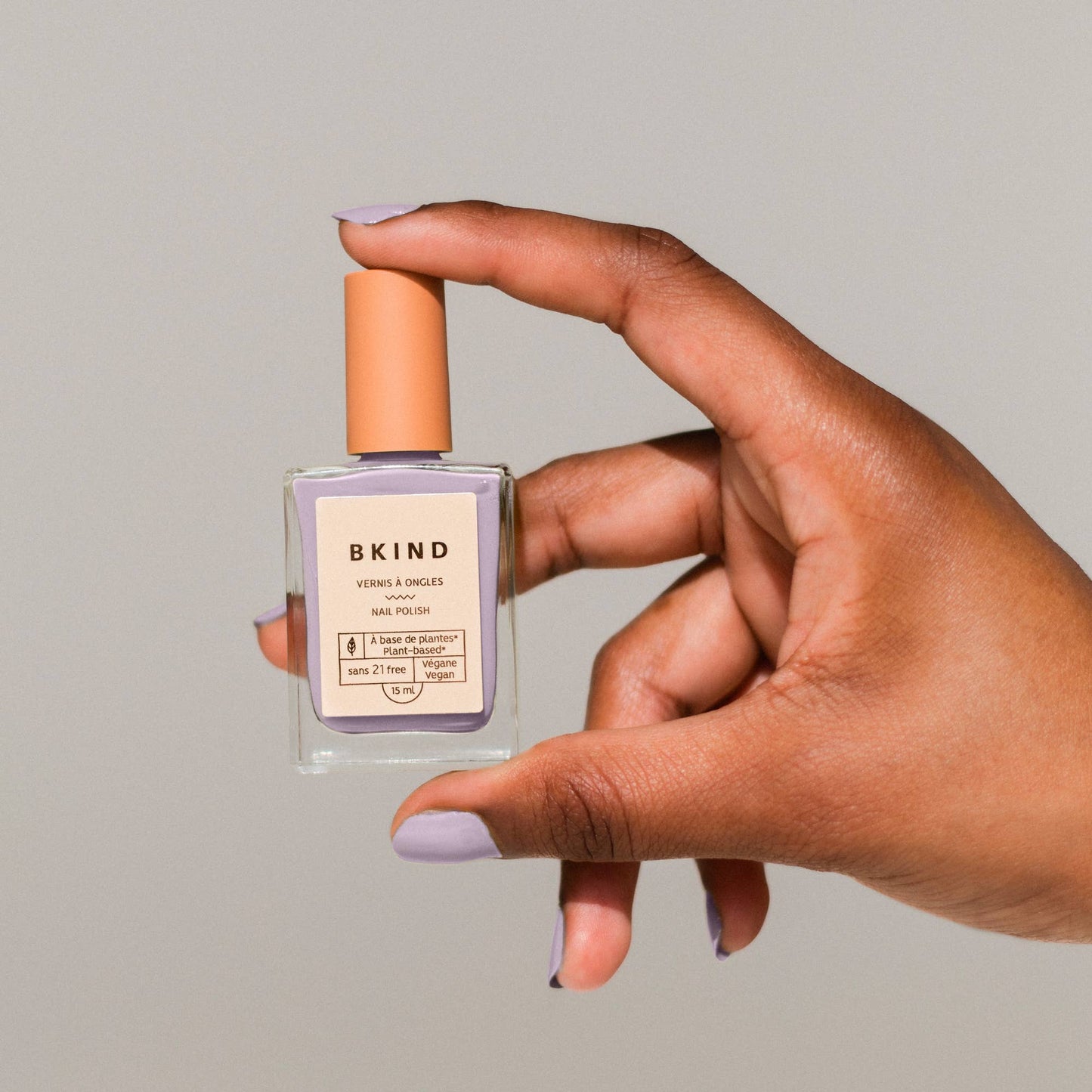 A hand holding a clear bottle of BKIND brand vegan nail polish in a light purple color, with a label and cap visible.