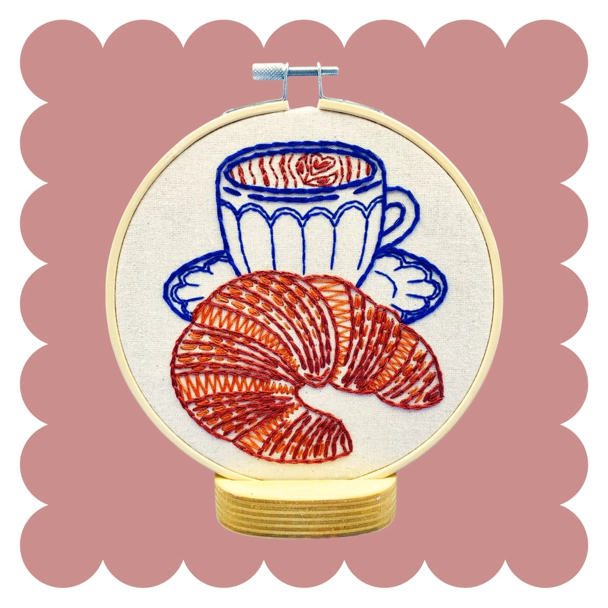 A circular embroidery hoop displaying a stitched design of a blue coffee cup and saucer with red croissant embroidery on a cream fabric background, set against a dusty pink scalloped backdrop.