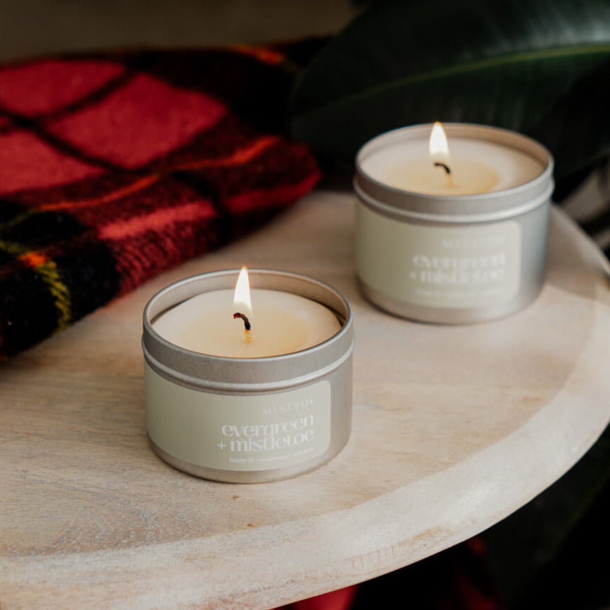 This image features the "Evergreen + Mistletoe Candle" from the Holiday Collection, thoughtfully designed to capture the essence of the festive season. The candle is shown in a warm, inviting setting, resting on a plaid fabric that enhances its cozy, seasonal appeal.