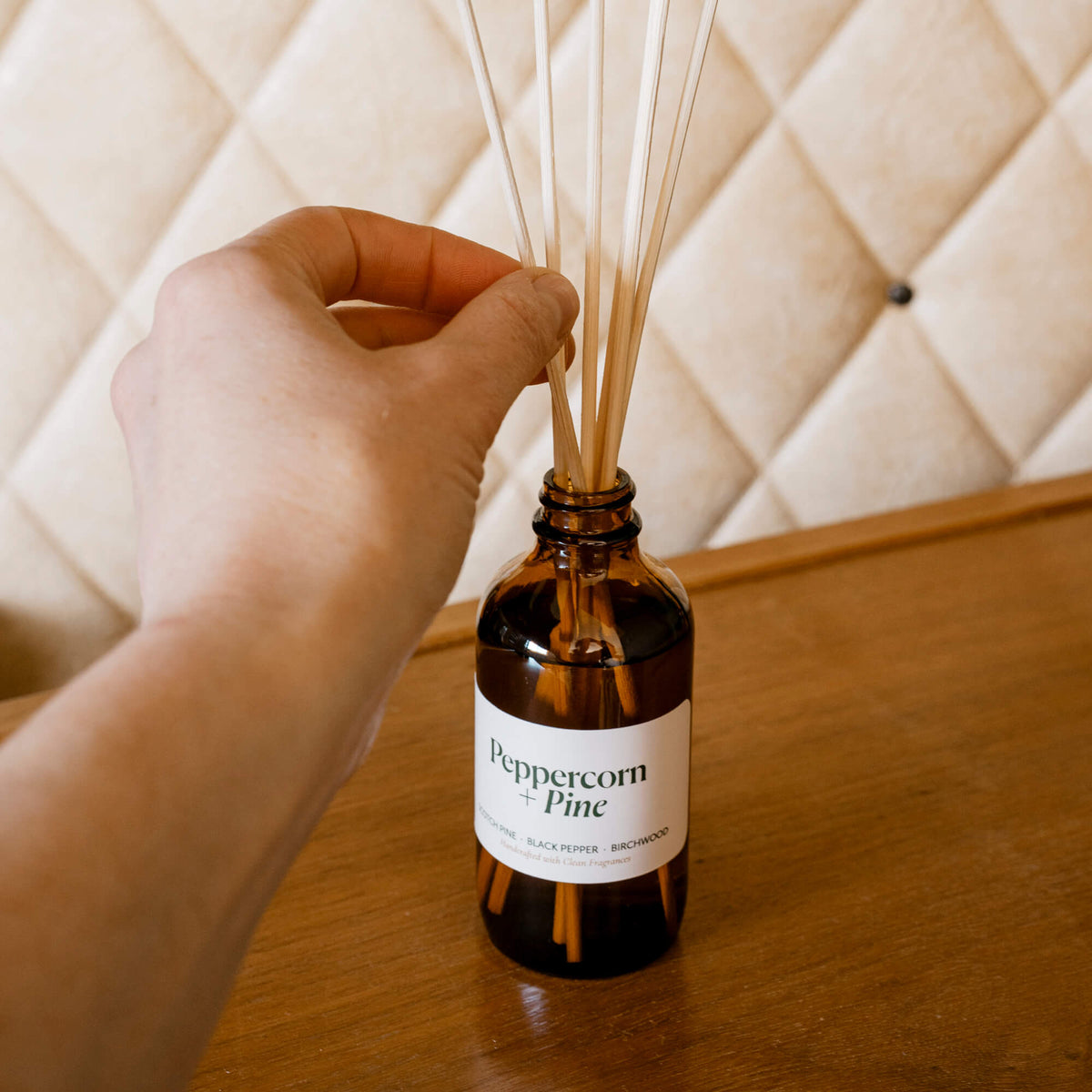 Hand arranging reeds in Misiyo Peppercorn + Pine reed diffuser on a wooden surface — easy setup, grounding pine & black pepper home fragrance, made in Canada