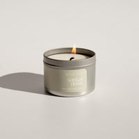 A Santal Dusk scented beeswax candle tin made in Edmonton, AB. The candle has scent notes of cedarwood, smoked vanilla and oud. The candle tin is silver with a sage green label and is shown burning with a white backdrop.